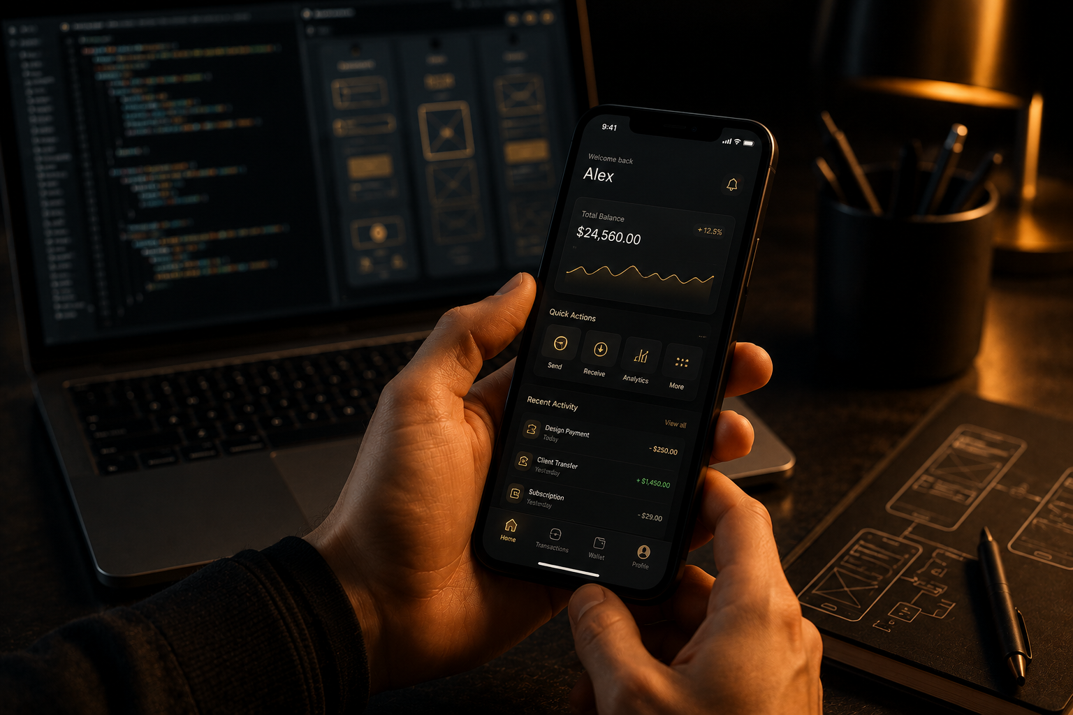 Hands holding a smartphone with a finance app UI, laptop with app wireframes in the background